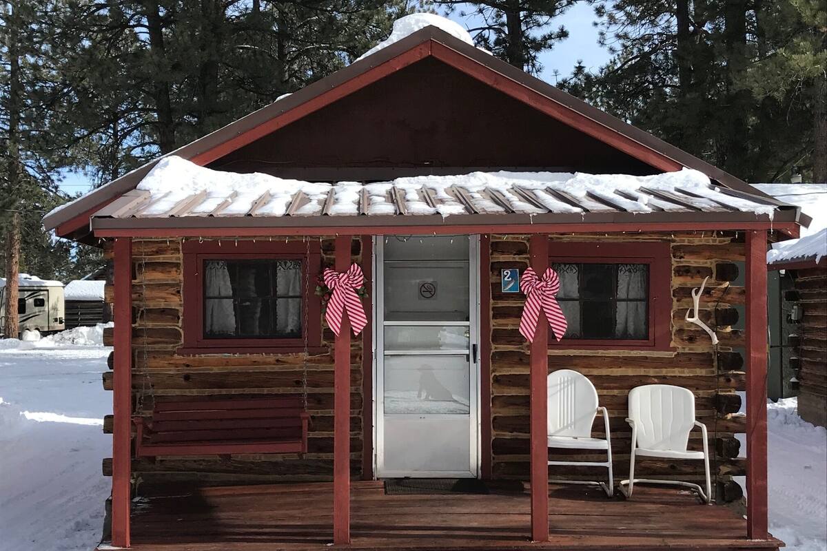 A log cabin with a front door, two windows, a porch with two white chairs, and a snow-covered roof.