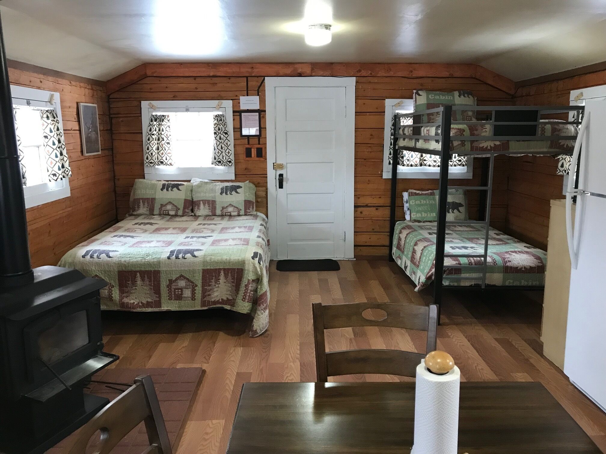 Una habitación acogedora estilo cabaña, con paredes de madera, una litera, una cama individual, una mesa con sillas, un televisor y una puerta.