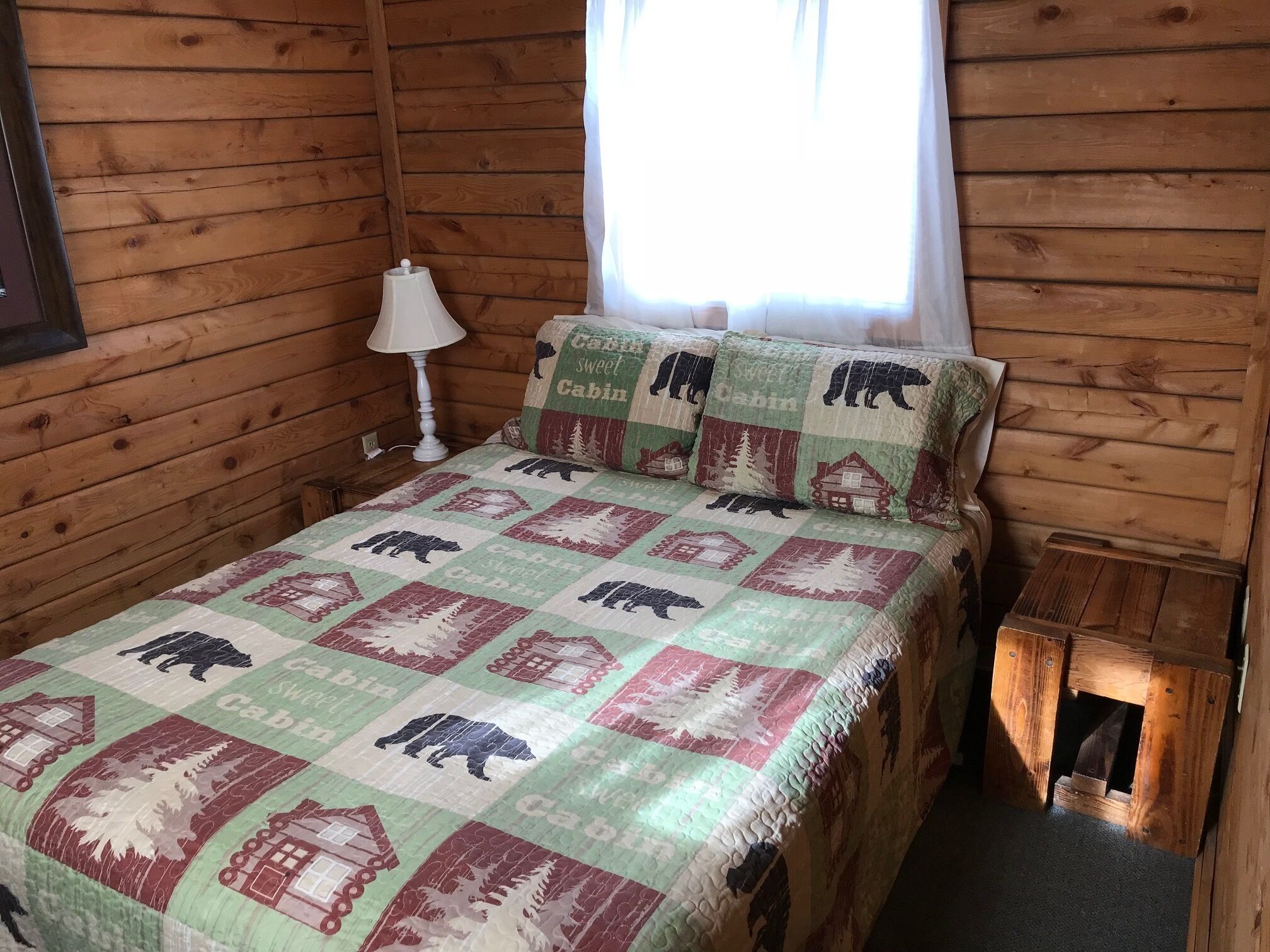 A wooden cabin-style room with a bed, a nightstand, a lamp, and a window with curtains.