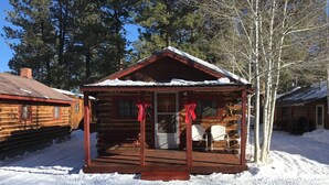 A log cabin with a porch, snow on the roof, and a red bow decoration.
