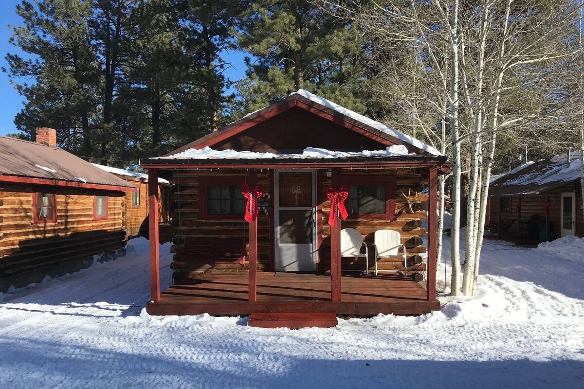 A log cabin with a porch, snow on the roof, and a red bow decoration.