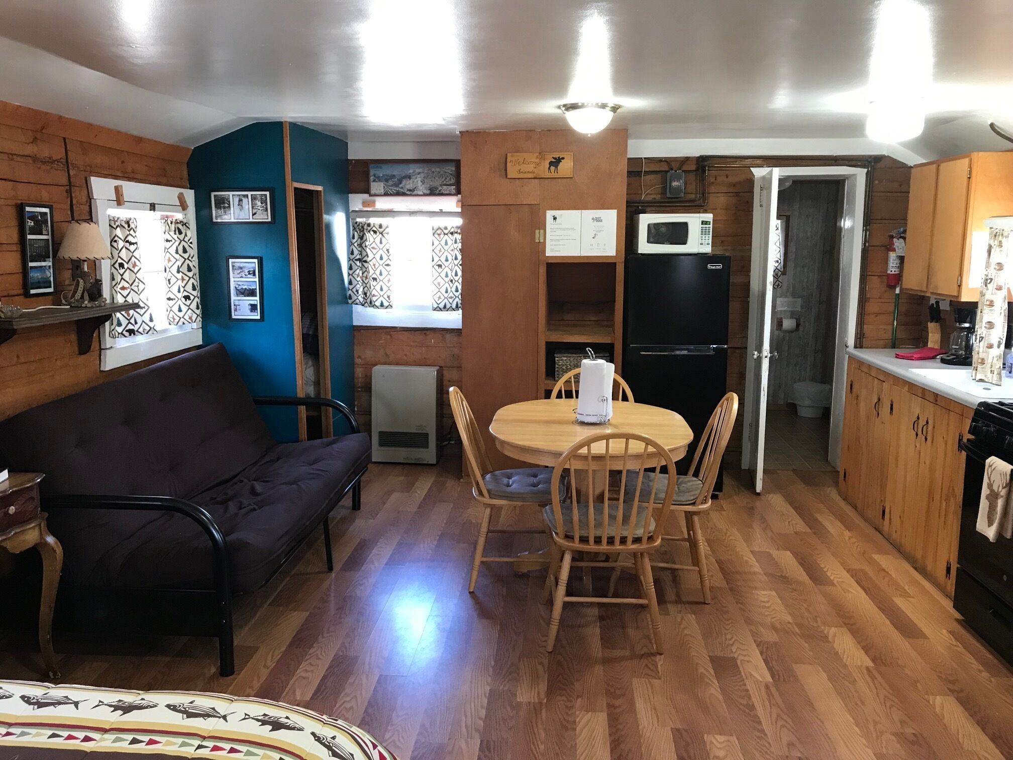 Een compact woongedeelte met een houten vloer, een eethoek met een ronde tafel en stoelen, een zwarte bank, een kleine kitchenette met een magnetron en koelkast, en een raam met gordijnen met dessin.