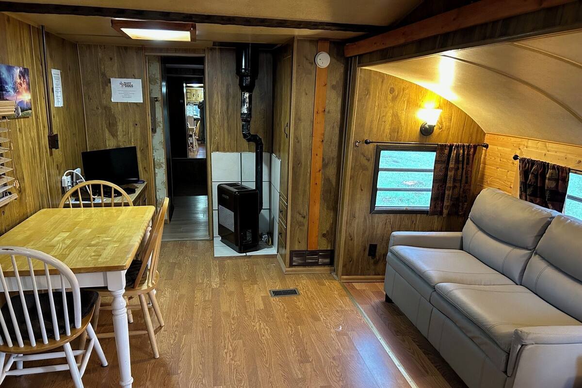 A cozy RV interior with a wooden dining table, white chairs, a gray sofa, and a television.