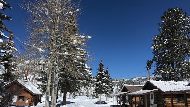 A snow-covered lodge with log cabins, a clear blue sky, and a snowmobile parked nearby.