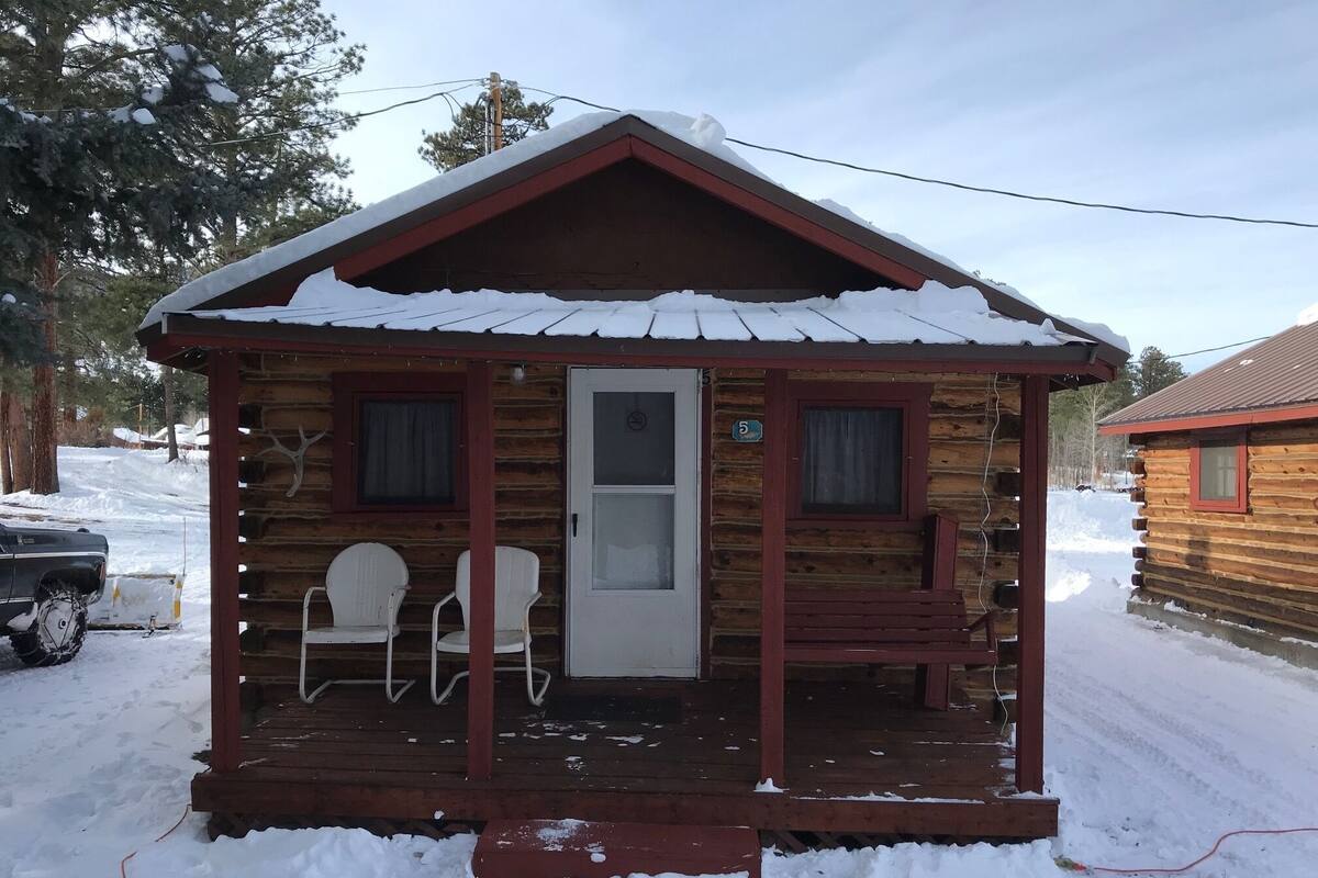 A small log cabin with a porch, two white chairs, and a bench.