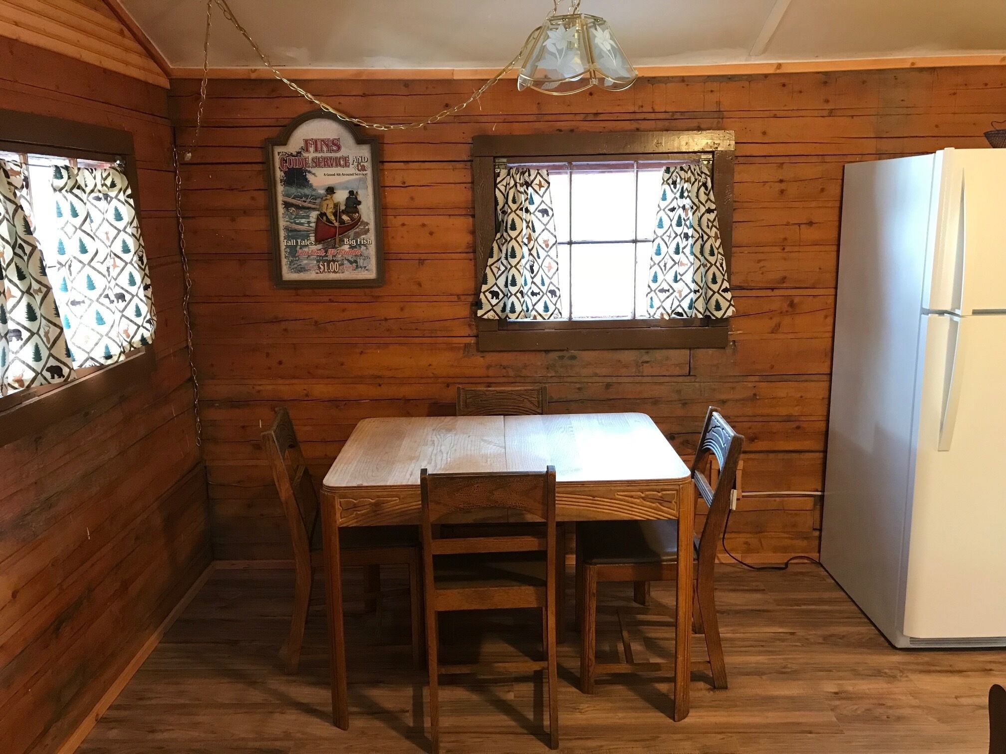 En stuga med träpanel på insidan, med ett matbord och stolar, ett kylskåp och fönster med ett mönstrat glas.