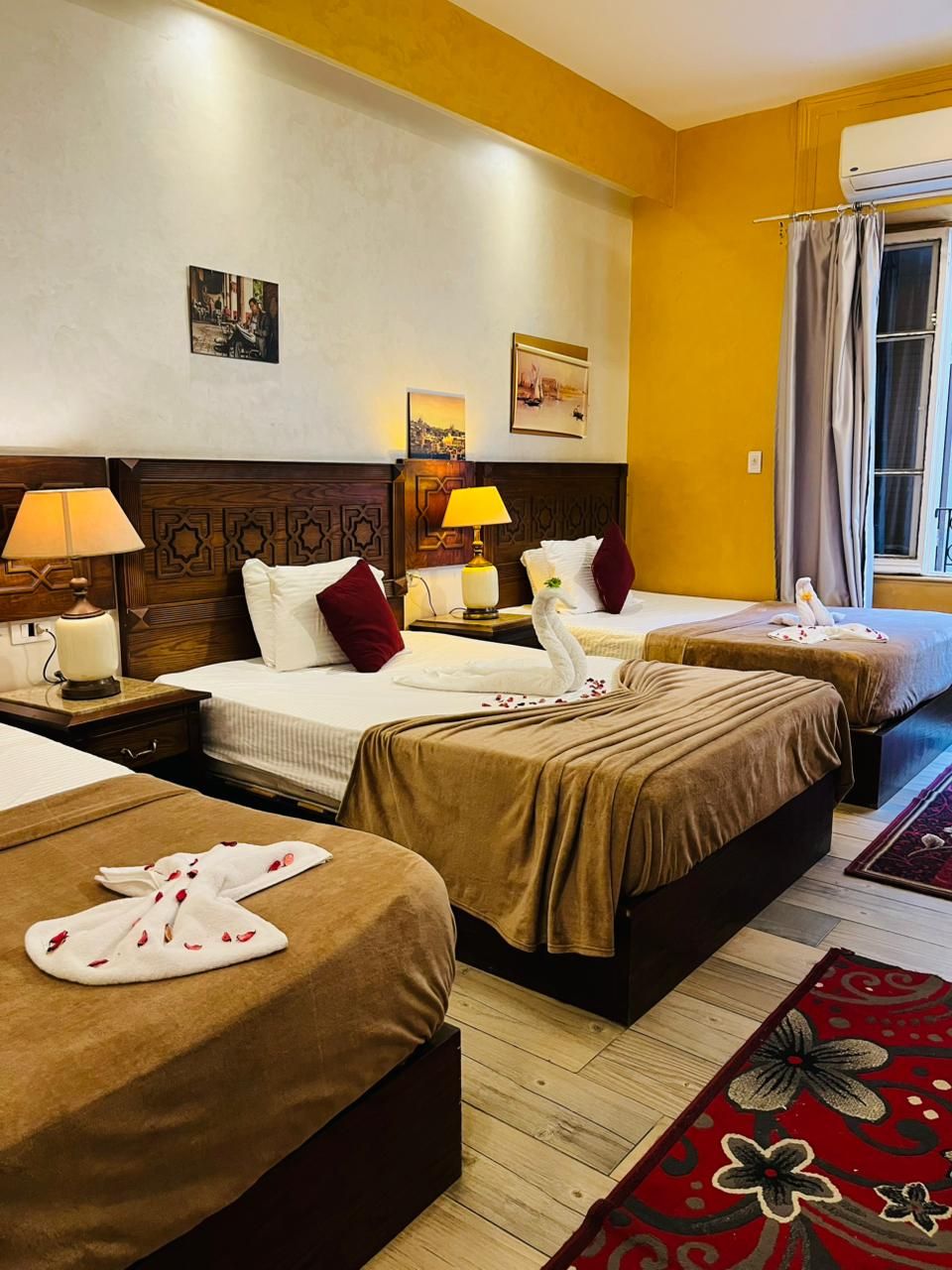 A hotel room with two beds, a wooden headboard, a nightstand with a lamp, and a window with curtains.
