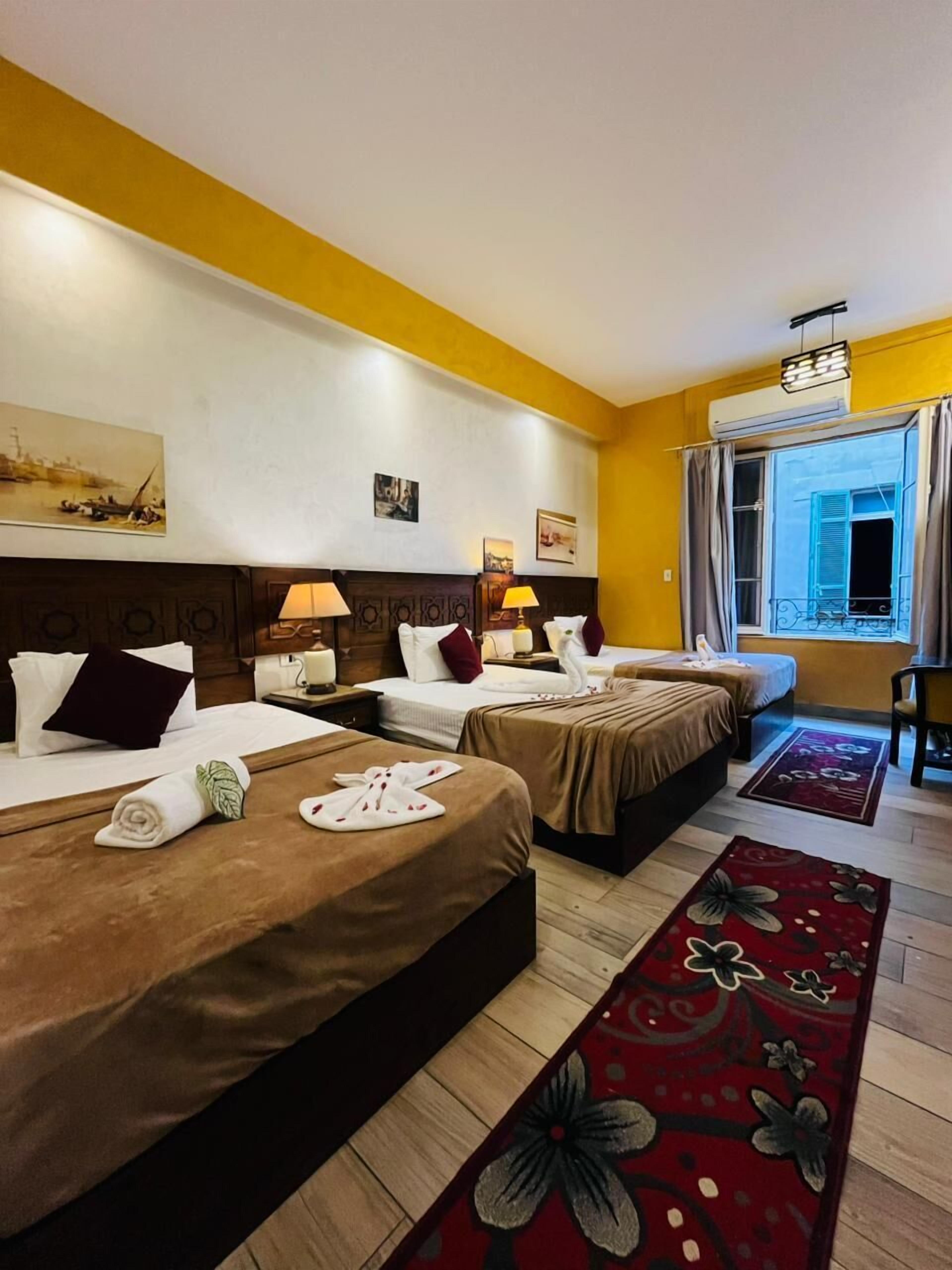 Habitación de hotel con dos camas, un ventanal con cortinas, un cuadro en la pared y un tapete floral rojo.