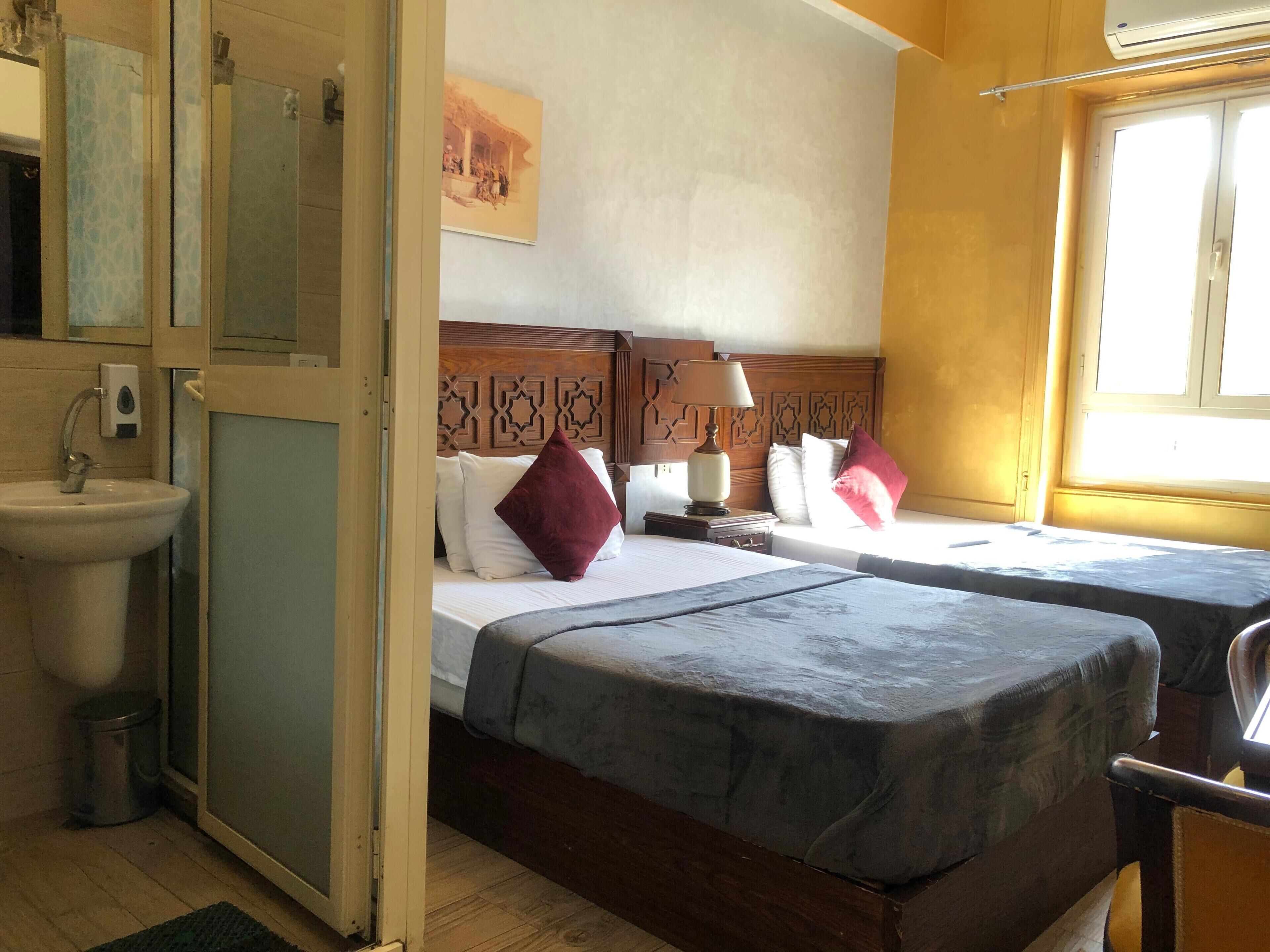 A hotel room with two beds, a bedside table with a lamp, a window, and a bathroom with a sink and mirror.