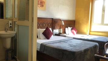 A hotel room with two beds, a bedside table with a lamp, a window, and a bathroom with a sink and mirror.