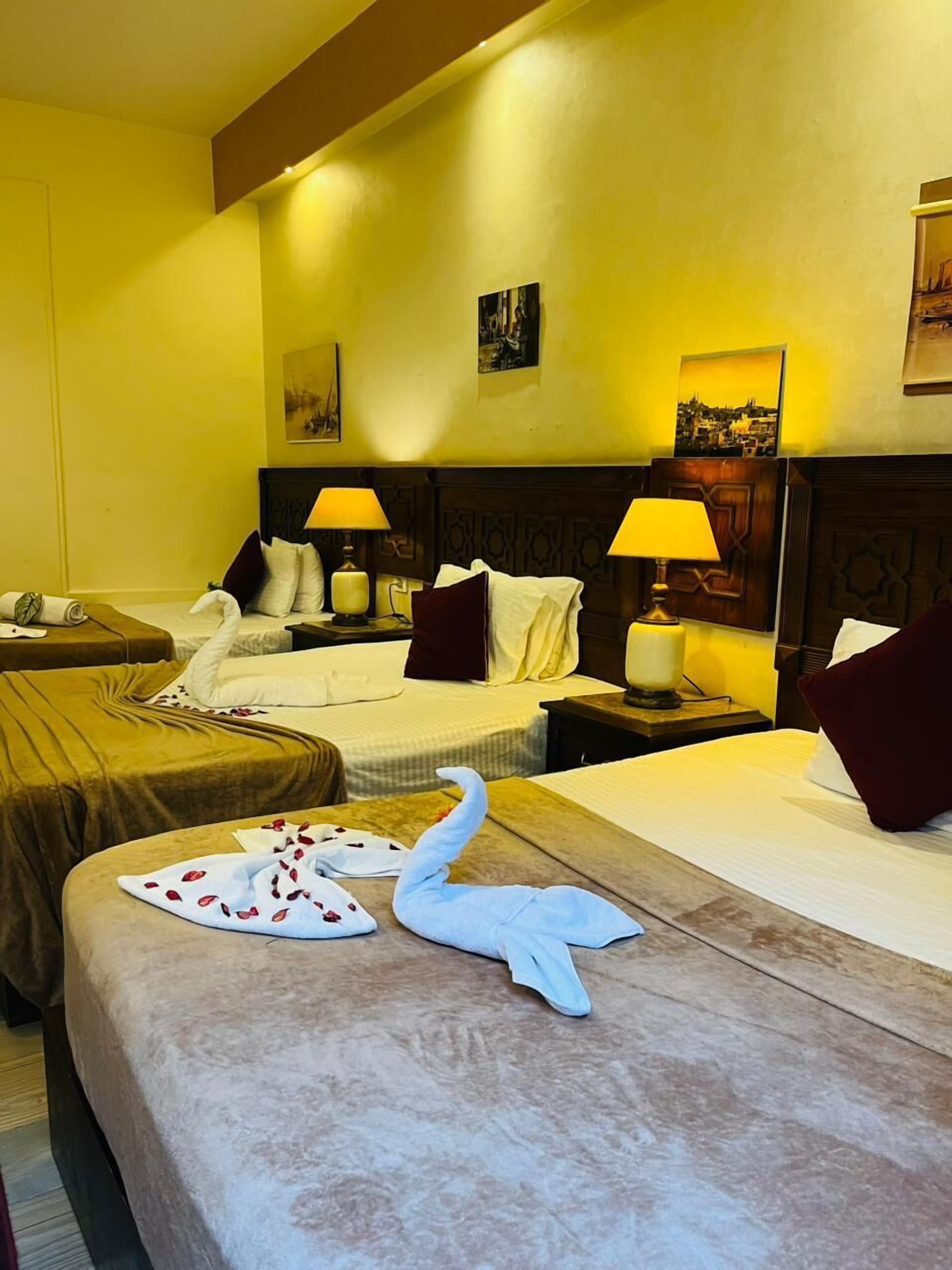 A hotel room with two beds, each with a headboard, bedside lamps, and pillows. There is a decorative swan on the bed in the foreground.