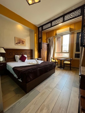 A hotel room with a large bed, a desk, and a chair. There is a window with curtains, a painting on the wall, and a wooden floor. - The Location Hotel (Cairo)