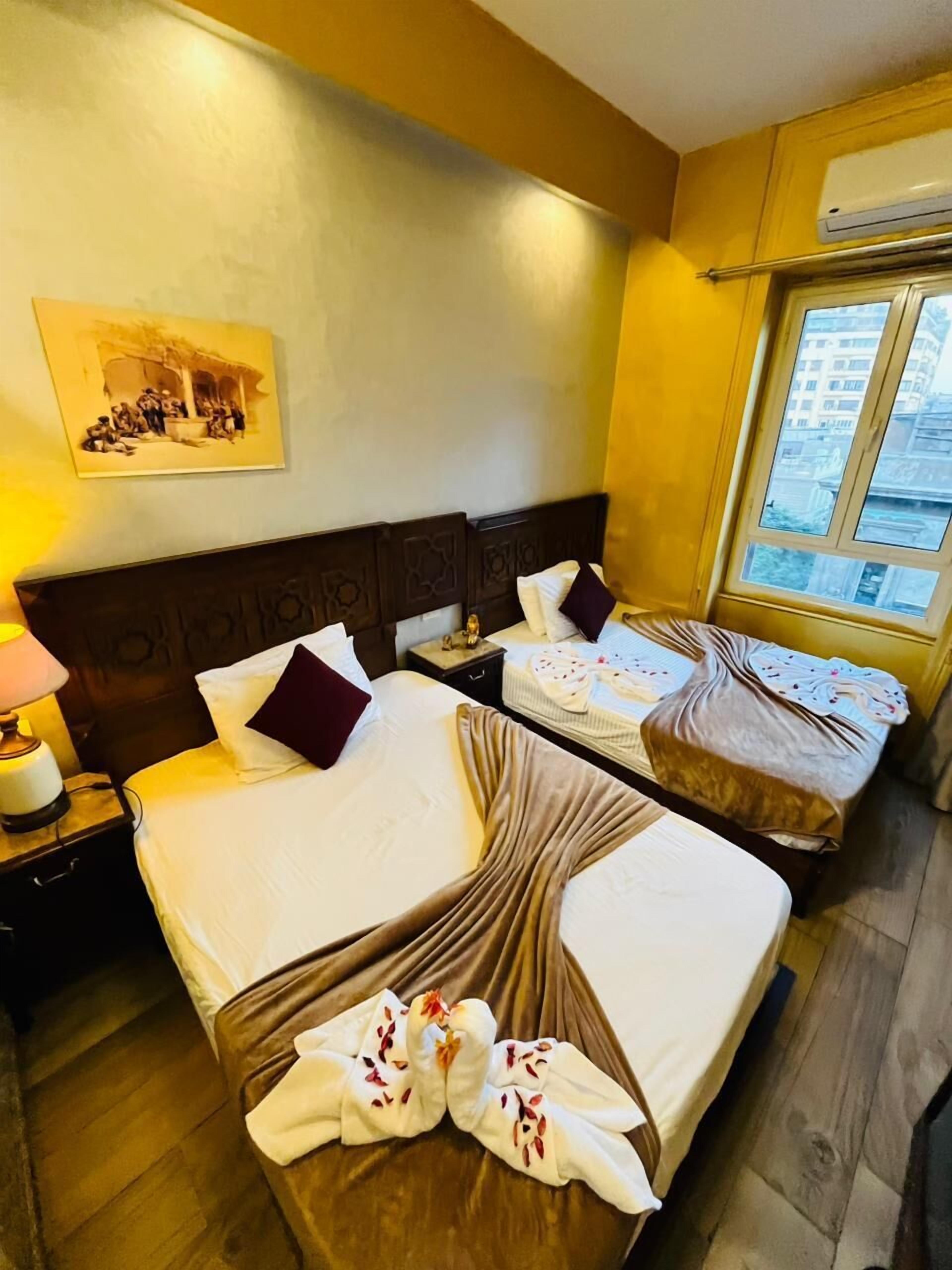 Quarto de hotel com duas camas, uma mesinha de cabeceira com abajur, um quadro na parede e uma janela com vista para edifícios.