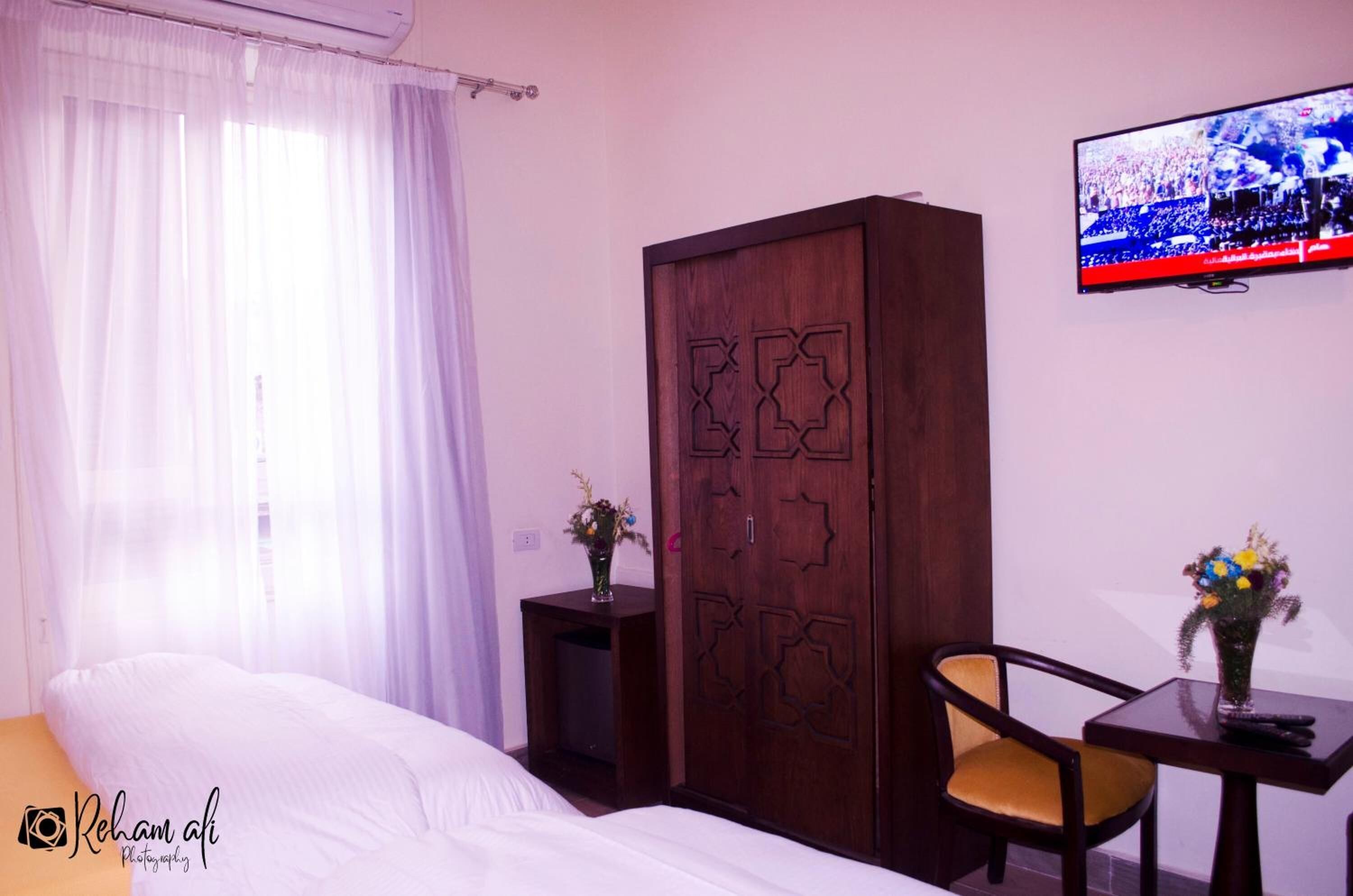Una habitación de hotel con una cama, un armario de madera, un televisor, una mesita con una silla y un jarrón con flores.