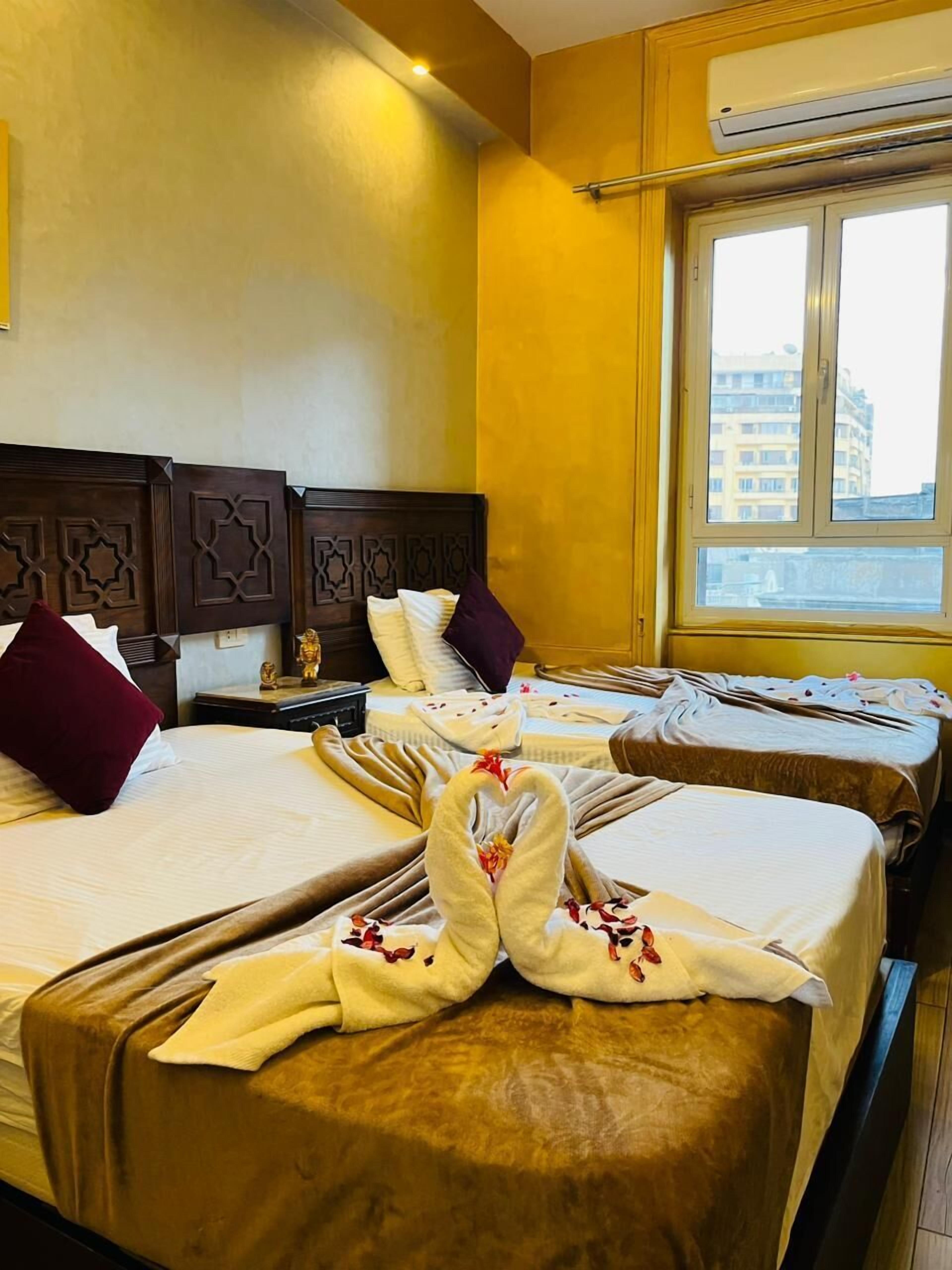 A hotel room with two beds, a wooden headboard with geometric patterns, a window with a view of buildings, and an air conditioning unit on the wall.