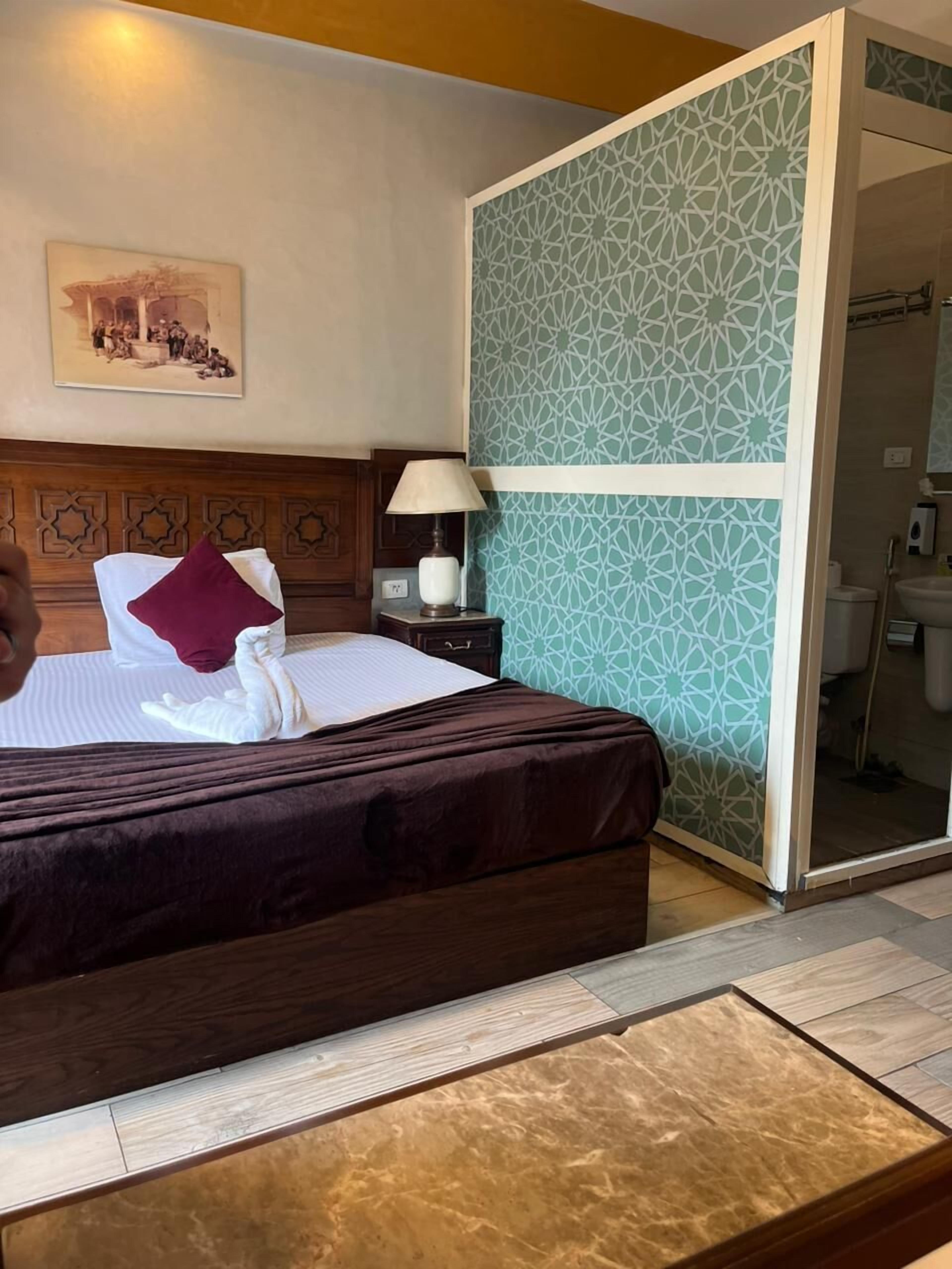 A neatly made bed with a brown headboard, a white pillow, and a red accent pillow. There is a nightstand with a lamp and a framed picture on the wall above the bed.