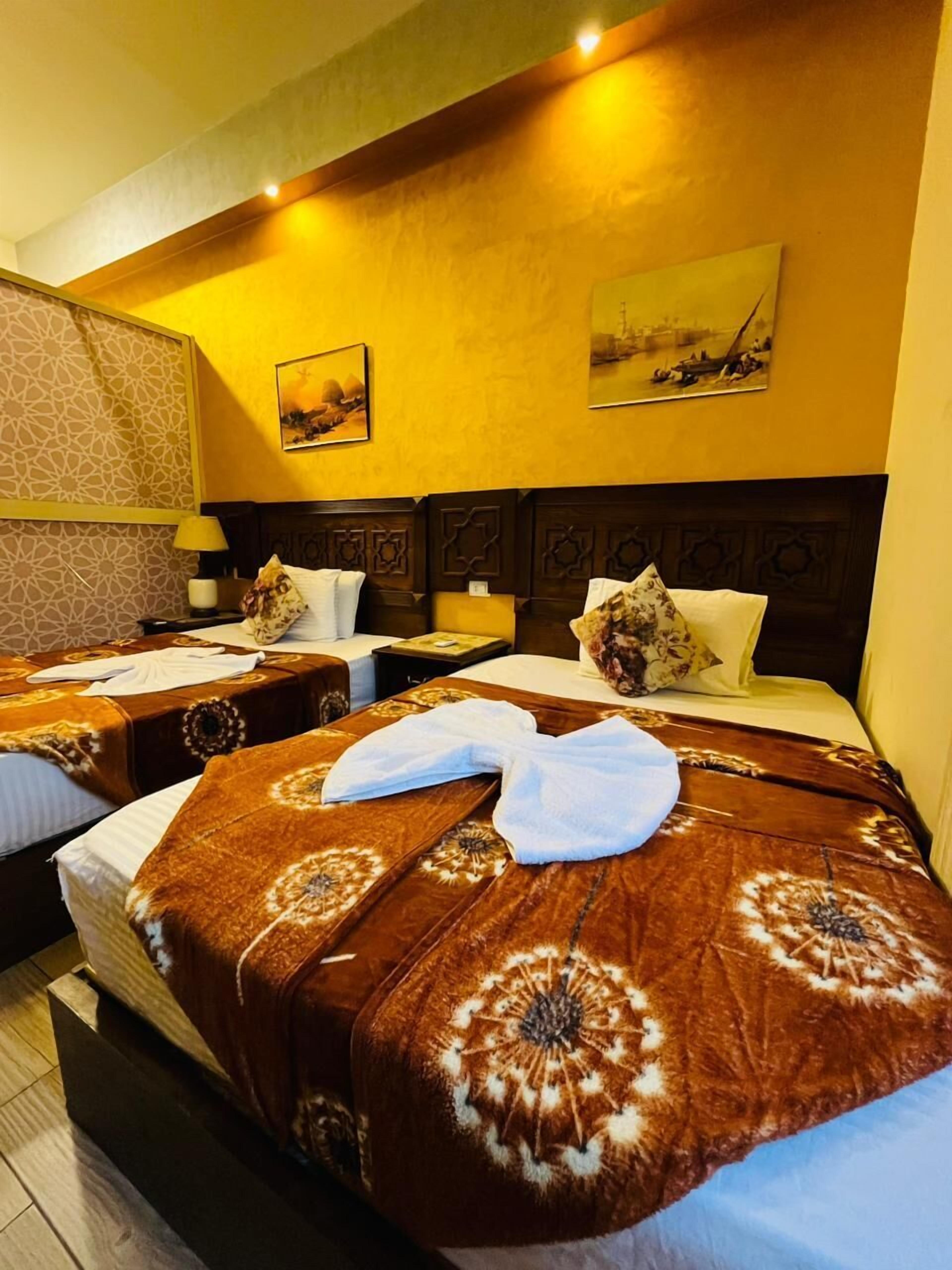 Habitación de hotel con dos camas, cada una con una colcha y almohadas estampadas. Hay lámparas de pared y un cuadro sobre las camas.