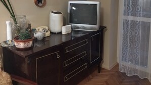 A room with a wooden cabinet, a television, a decorative wall piece, a potted plant, and a patterned curtain.