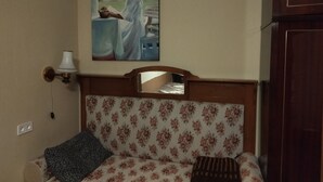 A bedroom with a floral-patterned bed, a painting of a woman reading, a wooden headboard, and a nightstand with a lamp.