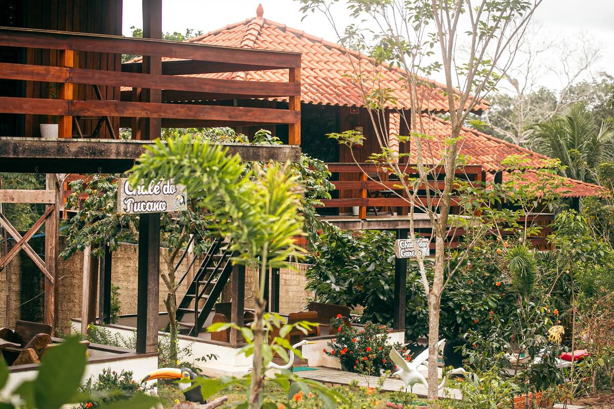A rustic wooden structure with a tiled roof, surrounded by lush greenery and a sign reading "Chá de Tucano".