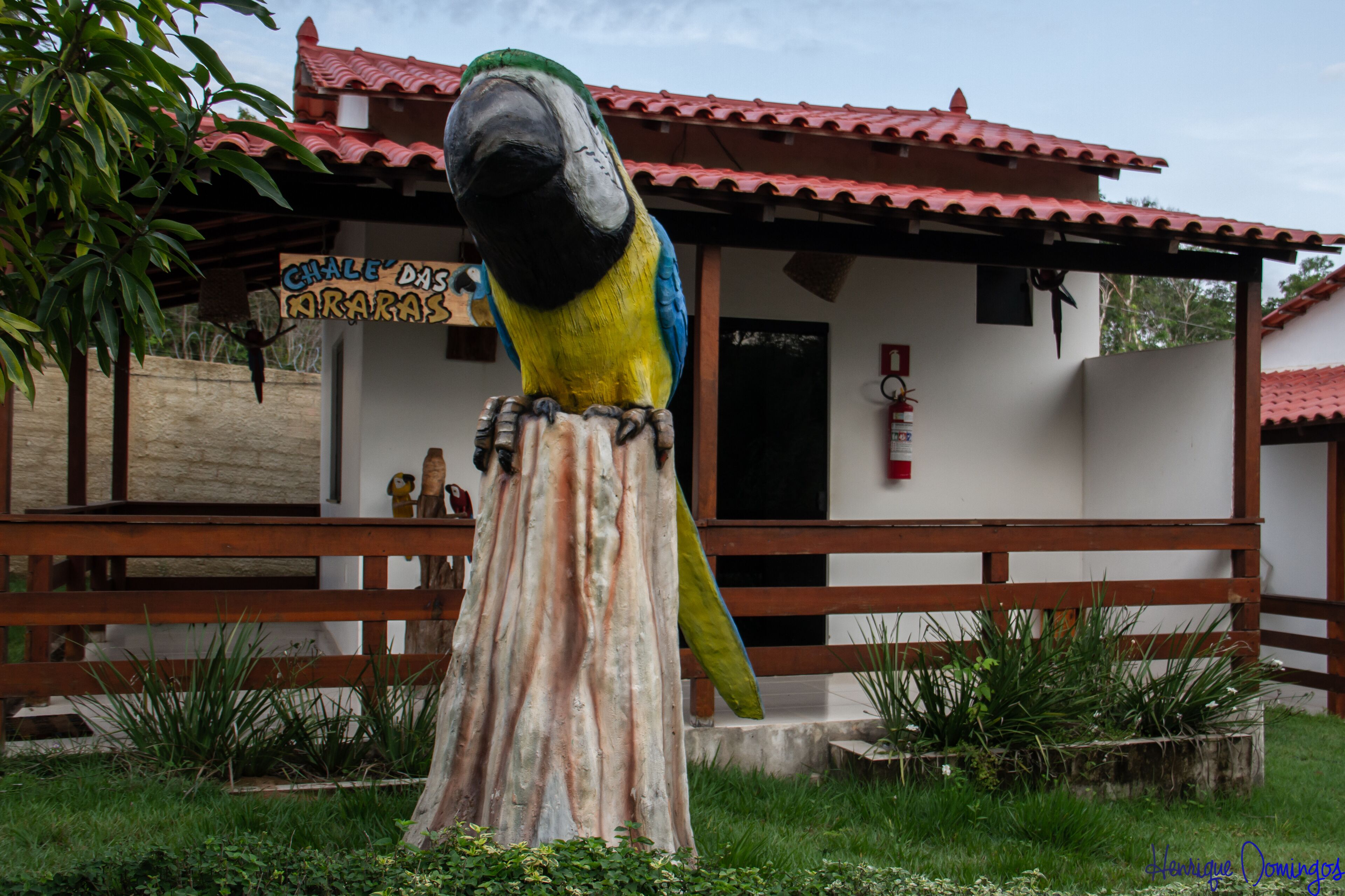 A large parrot statue in front of a building with a red-tiled roof and a sign that reads "Chalet Das Naranjas".