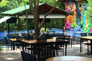 An outdoor dining area with wooden tables and chairs, a covered seating area, and a mural on the wall. - Pousada Amazônia (Santarém)
