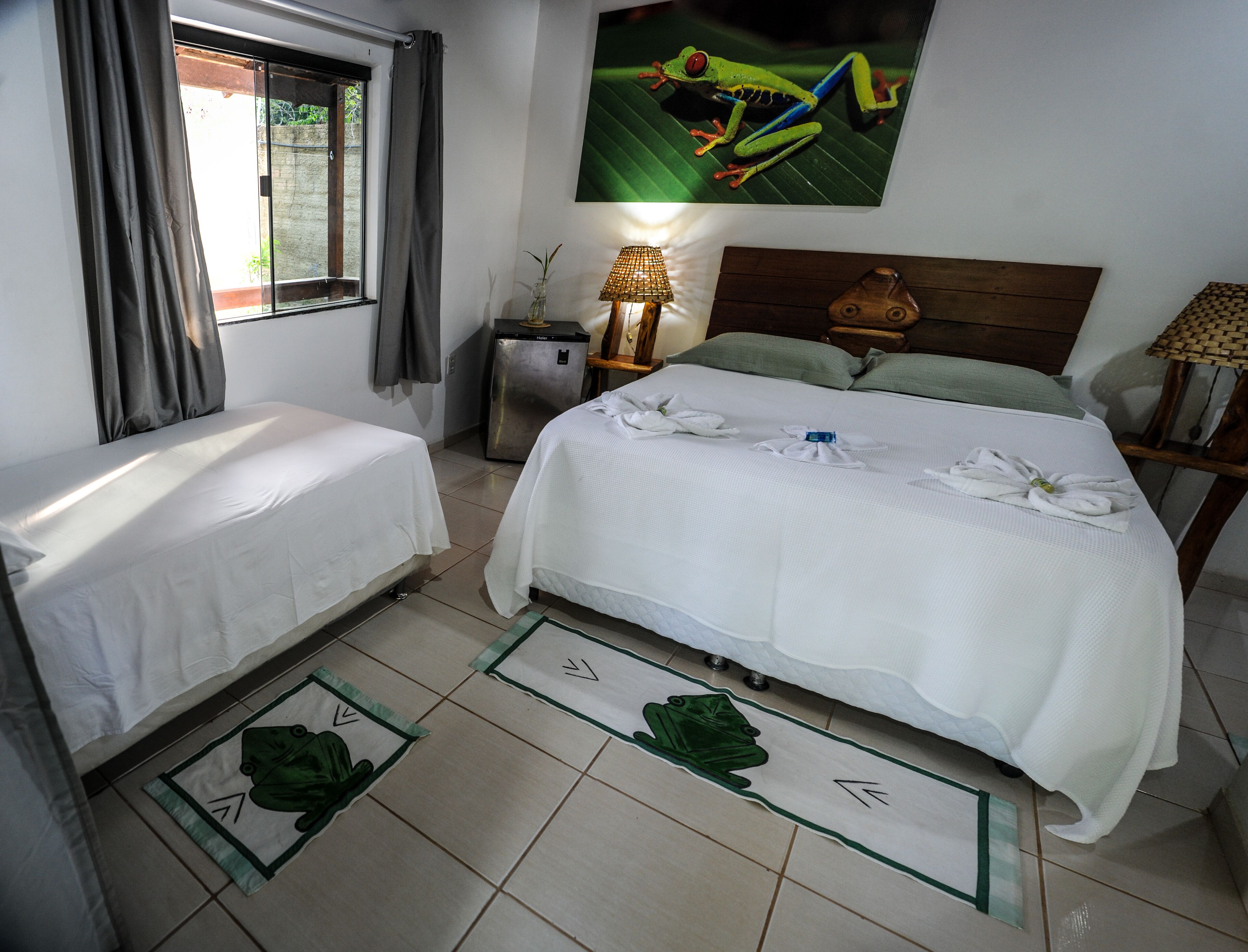 Habitación de hotel con dos camas individuales, una cama grande con toallas, un refrigerador pequeño, una lámpara, un cabecero de madera y un cuadro de una rana.