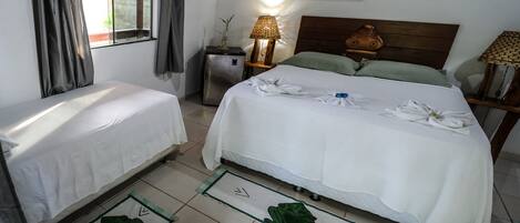 Habitación de hotel con dos camas individuales, una cama grande con toallas, un refrigerador pequeño, una lámpara, un cabecero de madera y un cuadro de una rana.