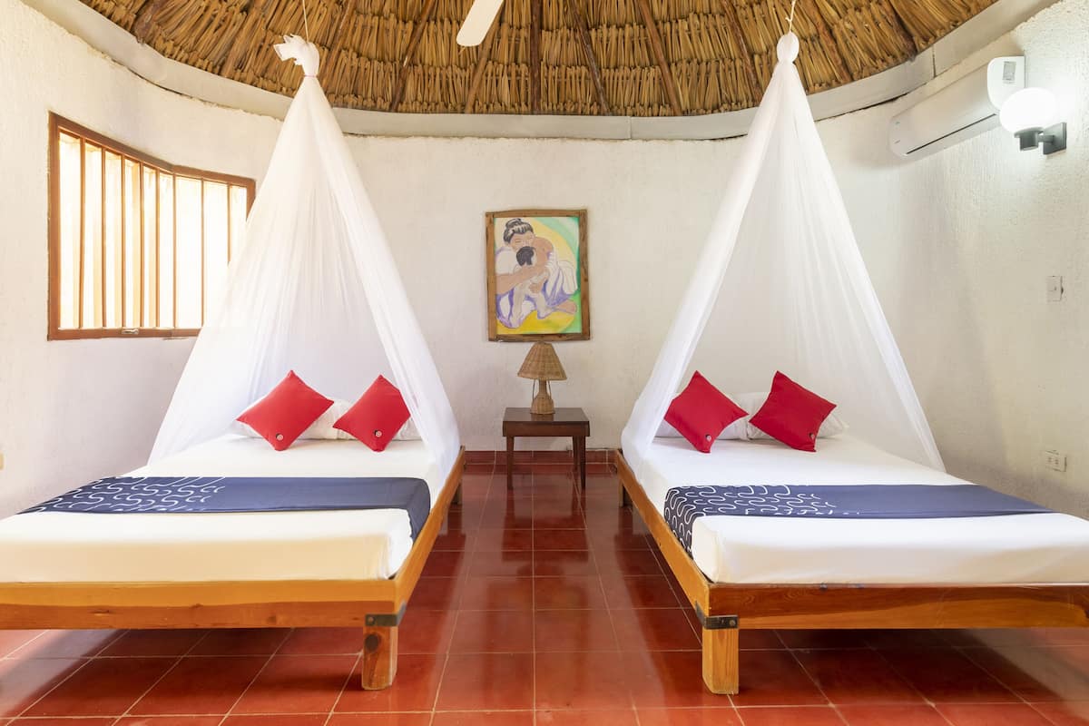 Ein Zimmer mit zwei Betten, jeweils mit Baldachin und roten Kissen, einem Deckenventilator, einem Gemälde an der Wand und einem kleinen Tisch.