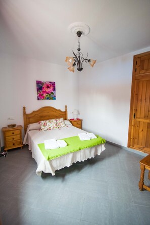 A bedroom with a wooden bed, a nightstand, a bedside table, a green throw blanket, and a floral painting on the wall.