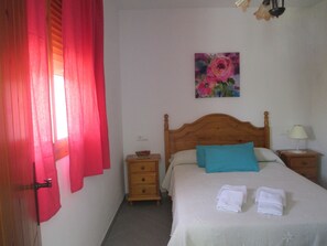 A bedroom with a wooden bed, a nightstand, a bedside table, a red curtain, and a floral painting on the wall.