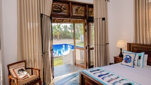 A bedroom with a bed, a chair, a nightstand, a lamp, and a view of a swimming pool. - Ardeen Villa (Kahandamodara)