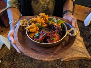 A person holding a bowl of food with a variety of items including a red sauce, vegetables, and fried items on a wooden tray. - Ardeen Villa (Kahandamodara)