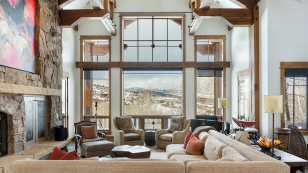 A spacious living room with a large window offering a view of a snowy landscape. There is a fireplace, comfortable seating, and a dining area with a table and chairs.
