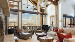 A spacious living room with a large window, wooden beams, a piano, and comfortable seating.
