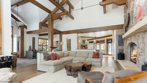 A spacious living room with a large sectional sofa, wooden beams, a stone fireplace, and a view of the outdoors.