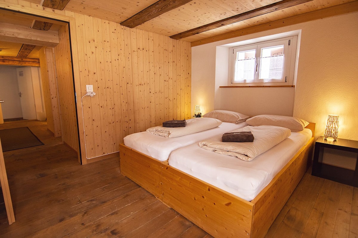 A wooden bed with two pillows and a blanket, a nightstand with a lamp, and a window with curtains.