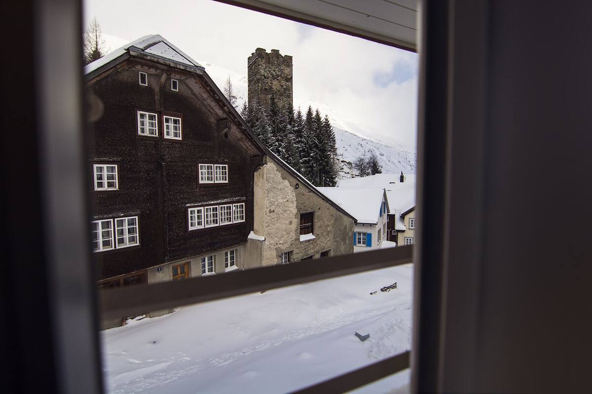 A traditional chalet with a stone chimney, multiple windows, and a snowy landscape visible through a window frame.