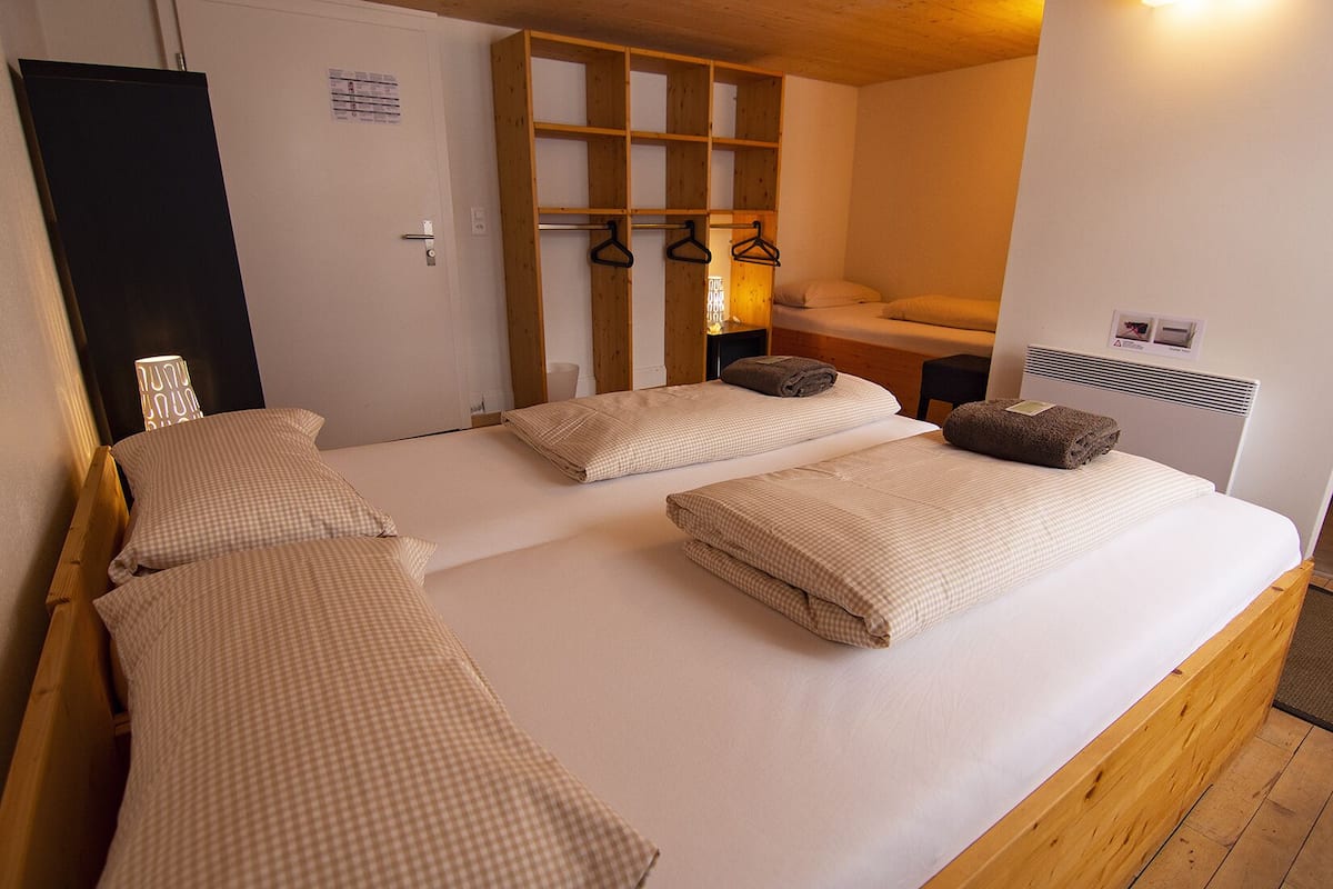 A room with two beds, wooden headboards, a hanging clothes rack, a bedside table, and a wall-mounted heater.