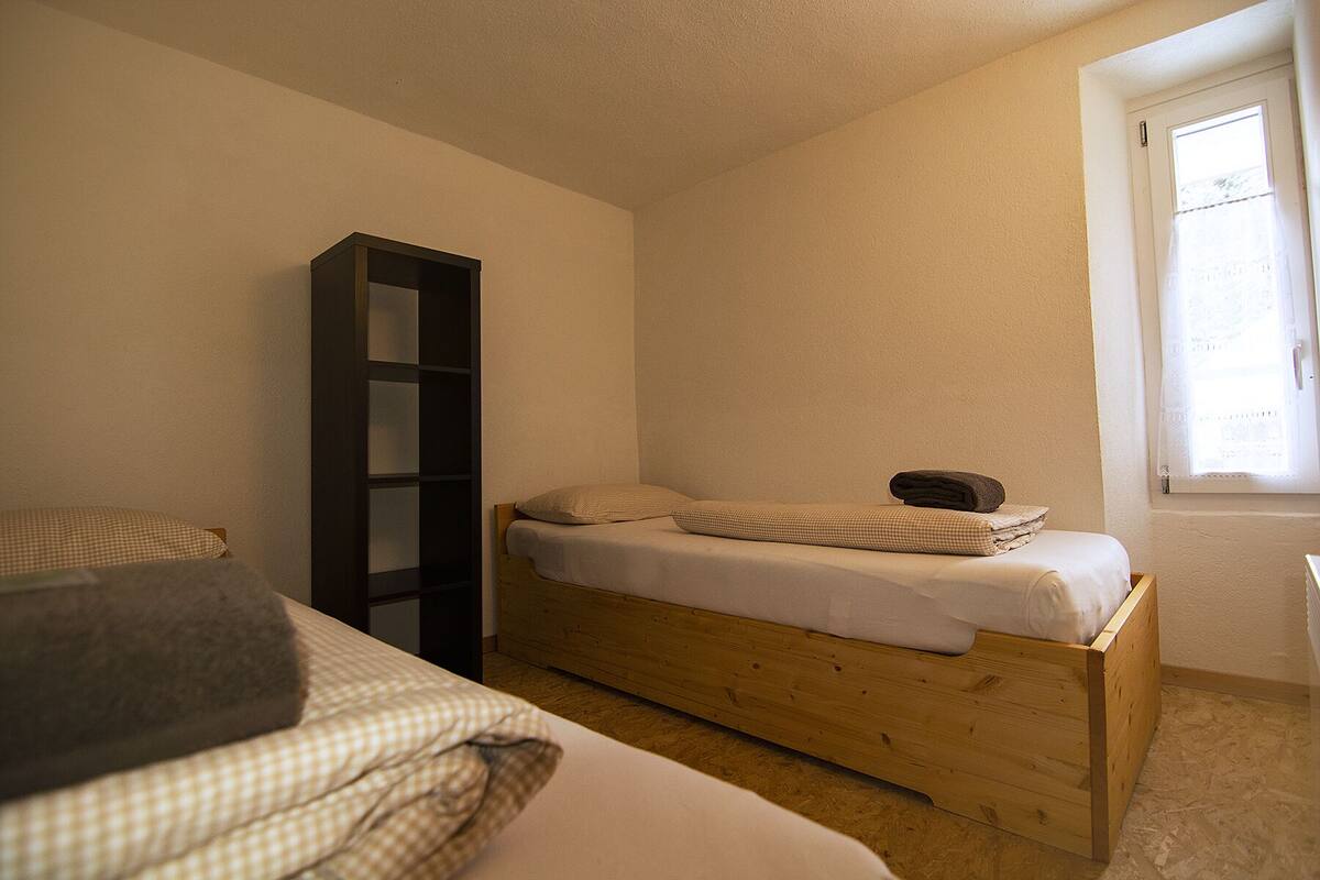 A small, single-bed room with a wooden bed frame, a tall bookshelf, and a window with a curtain.