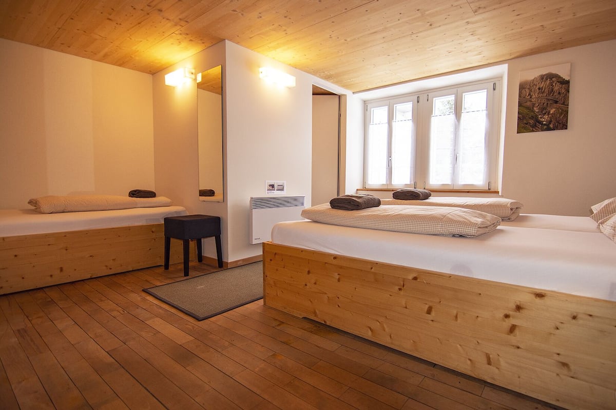 A cozy bedroom with two beds, wooden floors, a small stool, and a window with natural light.