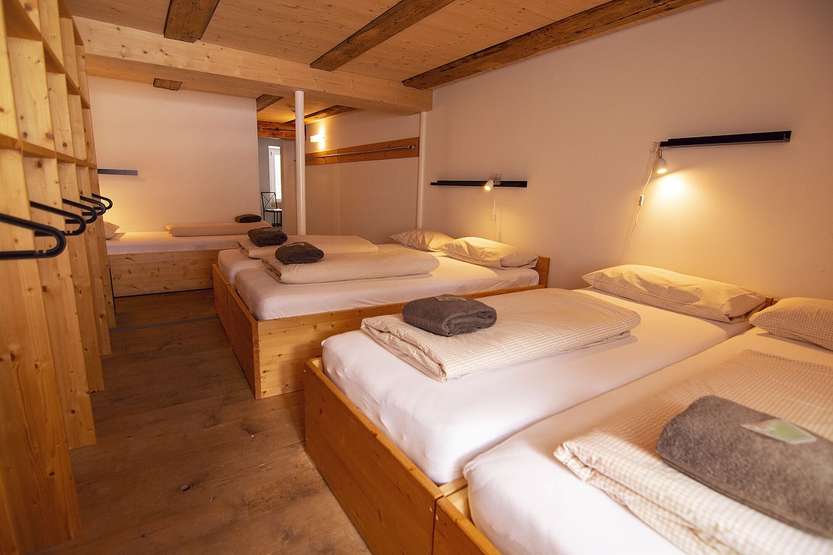 A room with three wooden beds, each with white linens and towels. There are wall-mounted lights above each bed.