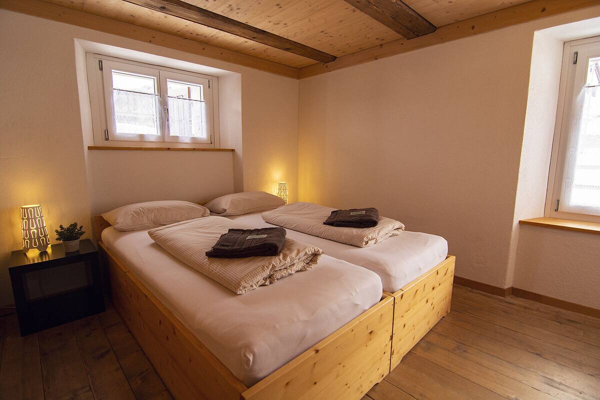 A double bed with a wooden frame, two bedside tables, a window with curtains, and a wall-mounted lamp.