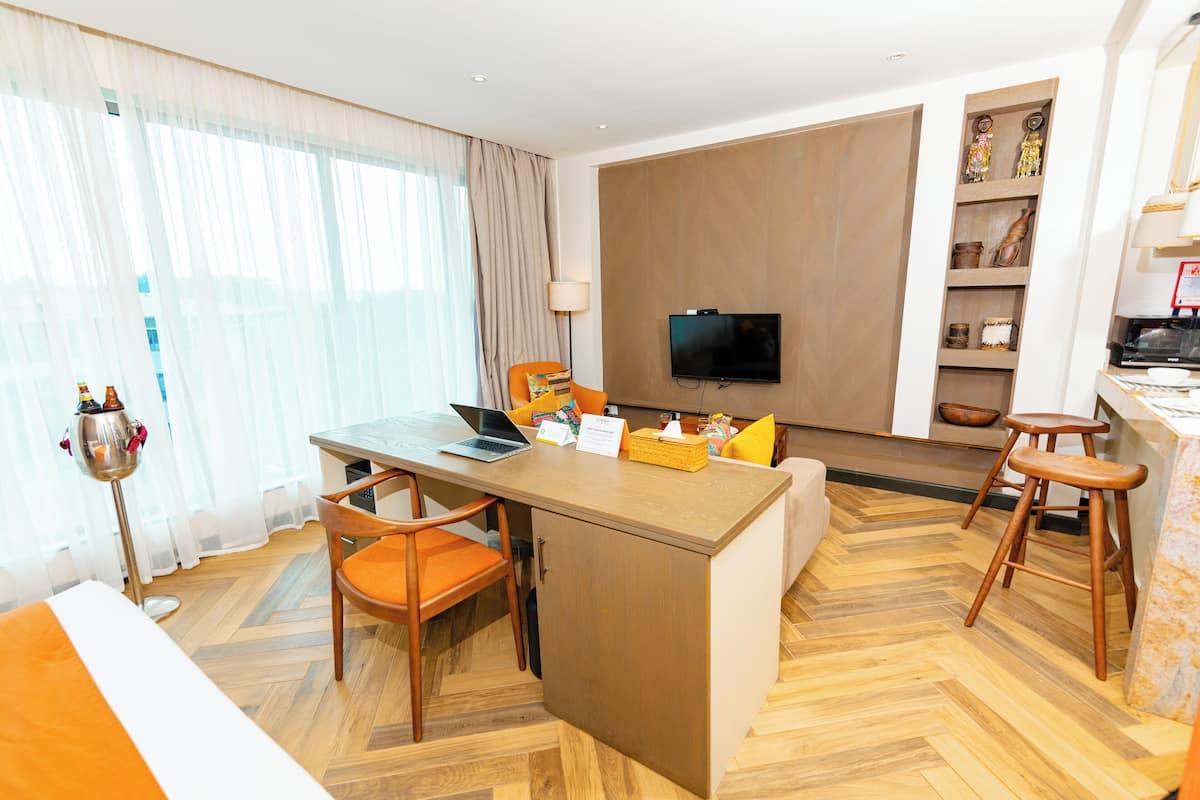 A hotel room with a wooden herringbone floor, a desk with a laptop, a television mounted on the wall, and a small kitchen area with stools.