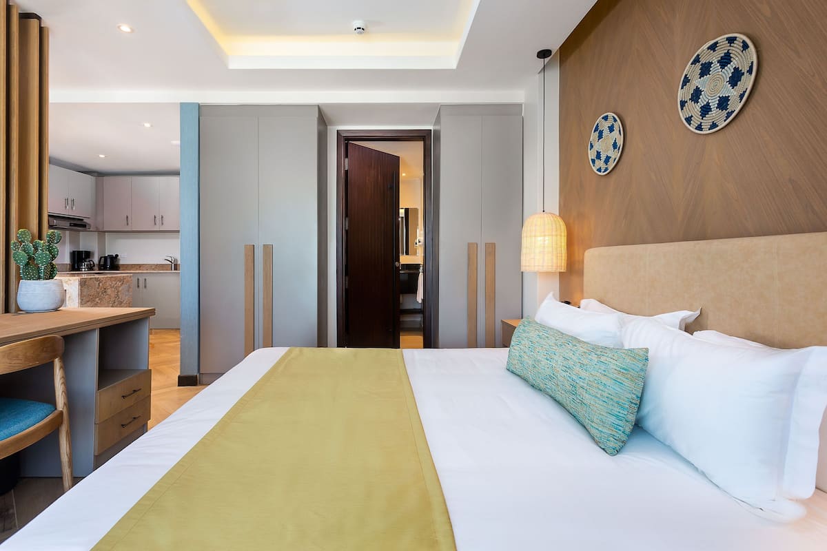 A modern hotel room with a bed, bedside table, lamp, and a view into the kitchen area.