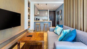 Ein modernes Wohnzimmer mit Flachbildfernseher, Couchtisch aus Holz, grauem Sofa mit blauen Kissen und einer Küchenzeile mit Barhocker.
