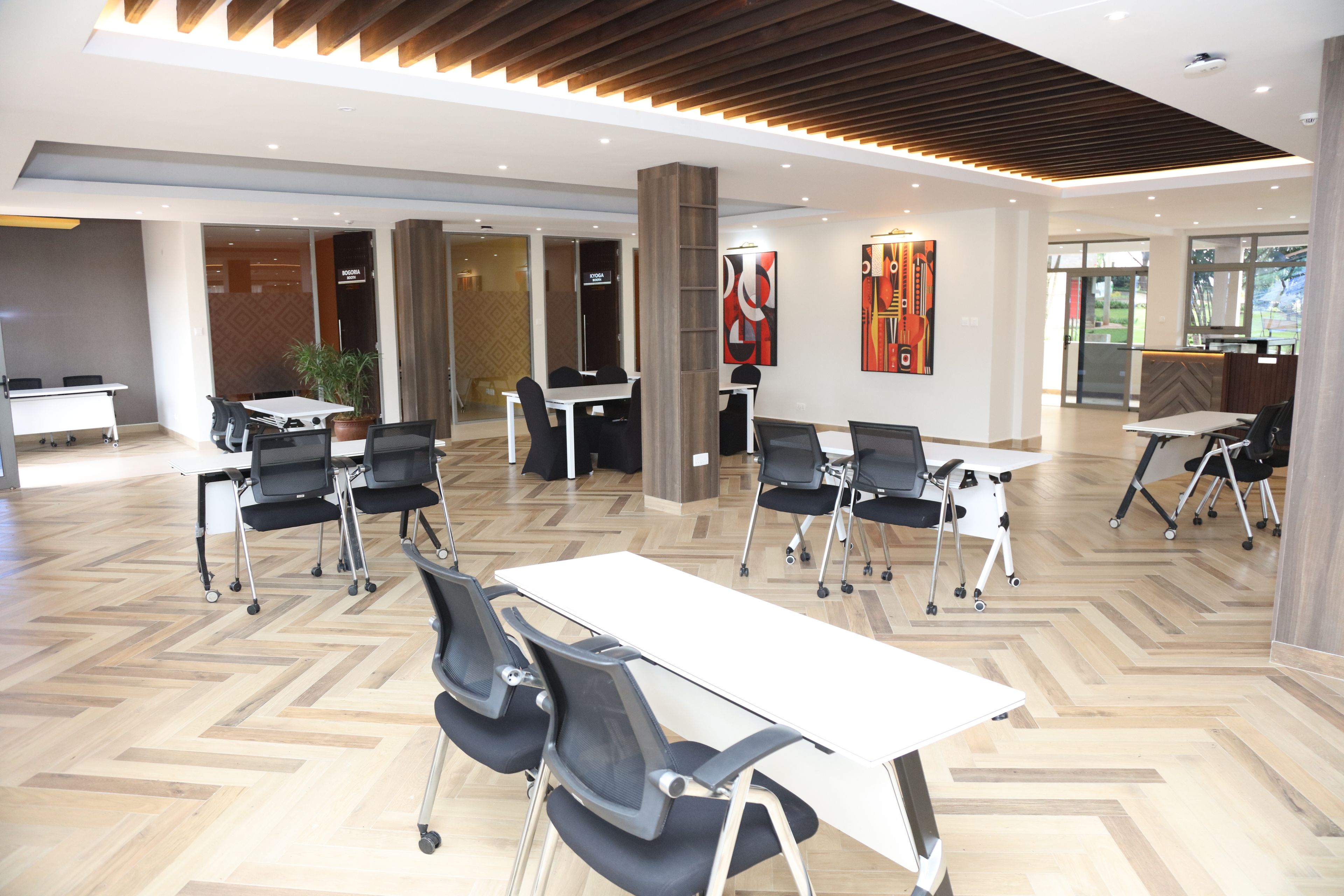 A modern conference room with white tables and black chairs, wooden flooring, and a high ceiling with recessed lighting.