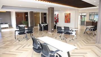 A modern conference room with white tables and black chairs, wooden flooring, and a high ceiling with recessed lighting.