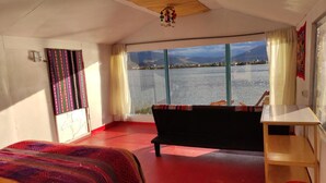 A room with a bed, a small table, a red floor, a window with a view of a lake and mountains, and a ceiling fan. - Uros Quechua Lodge (Puno)