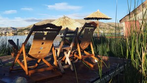 Two wooden lounge chairs with thatched umbrellas on a wooden deck overlooking a body of water and hills. - Uros Quechua Lodge (Puno)